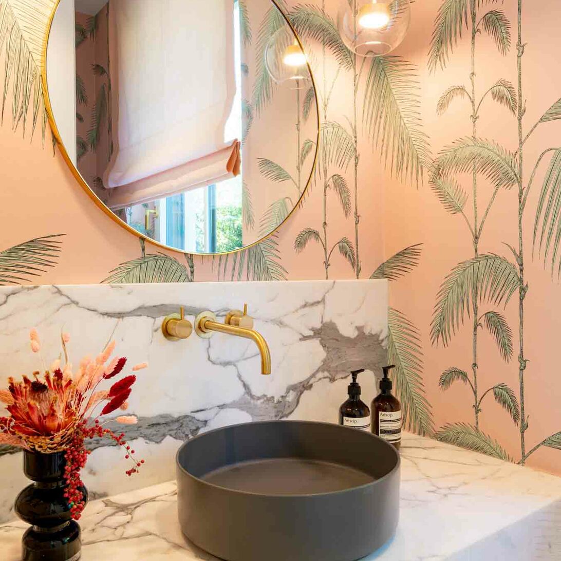 Ein stilvolles Bad mit Marmor-Waschtisch, dunklem Waschbecken, Palmen-Tapete, rundem Spiegel und goldenen Armaturen auf rosa Hintergrund. Ein stilvolles Bad mit Marmor-Waschtisch, dunklem Waschbecken, Palmen-Tapete, rundem Spiegel und goldenen Armaturen auf rosa Hintergrund.