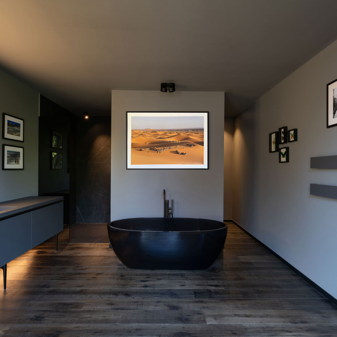 Modernes Badezimmer mit freistehender schwarzer Badewanne und Holzfußboden. Links ein beleuchteter Spiegel und rechts gerahmte Bilder an der Wand. Modernes Badezimmer mit freistehender schwarzer Badewanne und Holzfußboden. Links ein beleuchteter Spiegel und rechts gerahmte Bilder an der Wand.