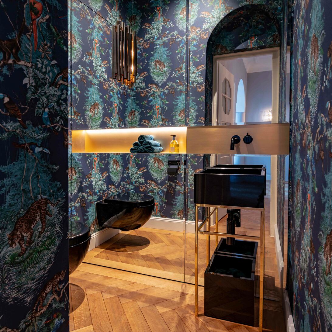 Dunkle, tropische Tapeten mit goldenen Akzenten, kombiniert mit eleganten schwarzen Armaturen und einem stilvollen Waschbecken. Der warme Holzfußboden rundet das edle Design ab.